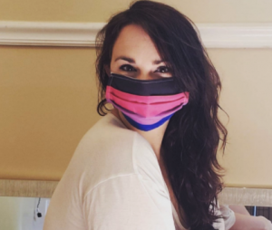 A woman with wavy brown hair looks to the camera. She's wearing a white shirt and a pink, purple, blue, and black face mask.