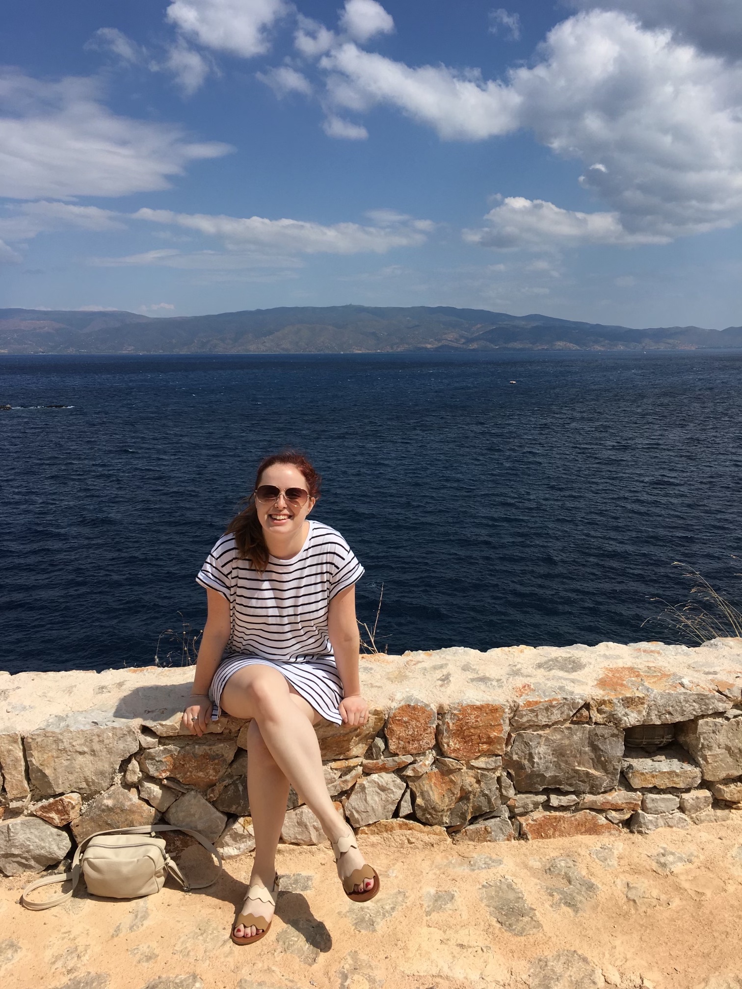 Patient Advocate Emily Byrn sits on a limestone ledge, smiling at the camera with the ocean in the background.